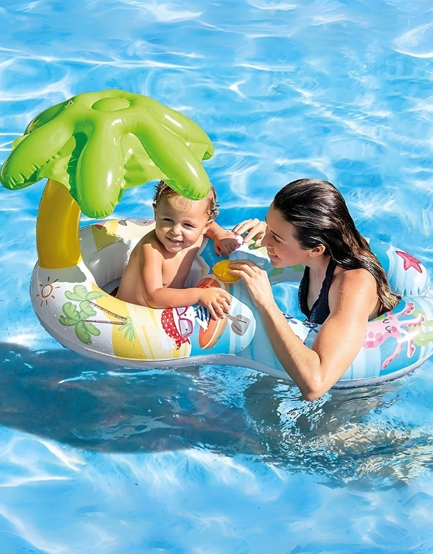 A smiling toddler sitting in an inflatable swim float with a palm tree canopy, accompanied by a woman in a pool.